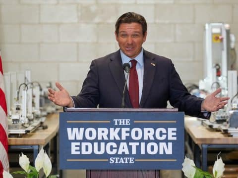 Man in a suit giving a speech at a podium reading 'The Workforce Education State' with an American flag on the left.