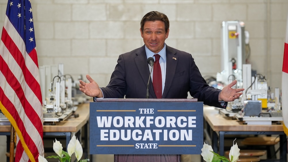 Man in a suit giving a speech at a podium reading 'The Workforce Education State' with an American flag on the left.