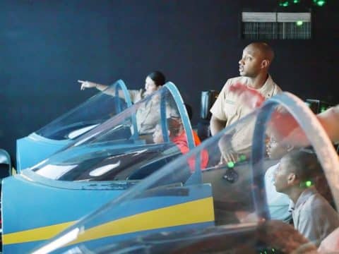 Group of people in beige uniforms seated in blue flight-simulator cockpits with clear canopies in a training room.