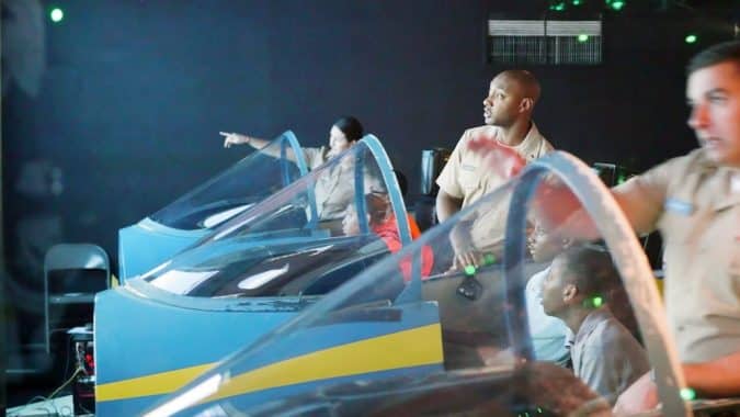 Group of people in beige uniforms seated in blue flight-simulator cockpits with clear canopies in a training room.