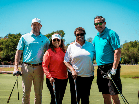 Four people posing on a sunny golf course, each holding a golf club and smiling at the camera.