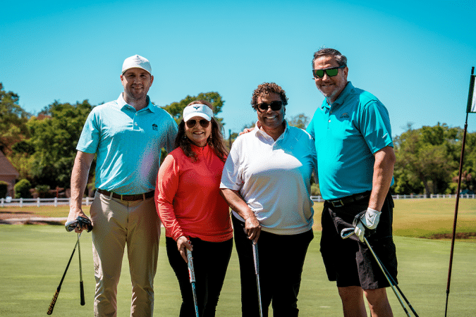 Four people posing on a sunny golf course, each holding a golf club and smiling at the camera.
