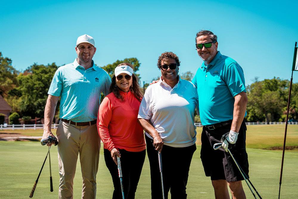 Four people posing on a sunny golf course, each holding a golf club and smiling at the camera.