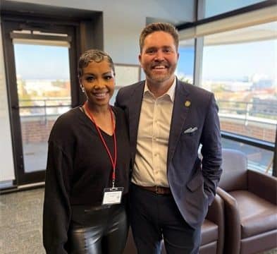 Two people posing together in a modern office lobby, woman with a badge on the left and man in a navy suit on the right, both smiling.