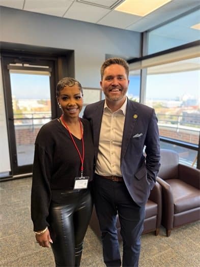 Two people posing together in a modern office lobby, woman with a badge on the left and man in a navy suit on the right, both smiling.
