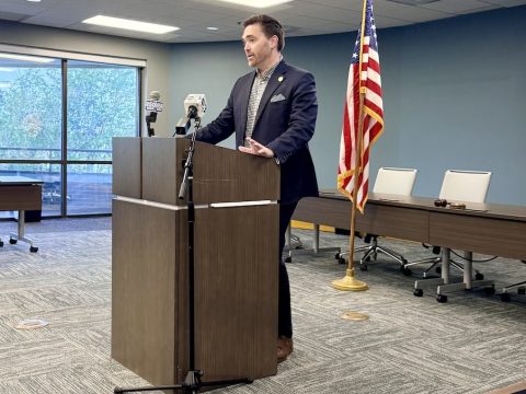 Man in a dark suit speaks at a wooden podium in a conference room with microphones and an American flag behind him.