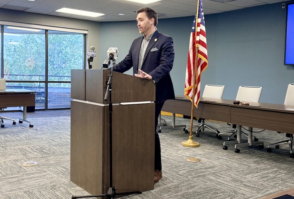 Man in a dark suit speaks at a wooden podium in a conference room with microphones and an American flag behind him.