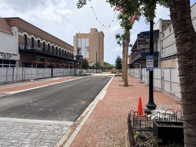 Urban street under construction with fenced sidewalk and brick path on the right, palm trees, and a bus stop sign in the distance.