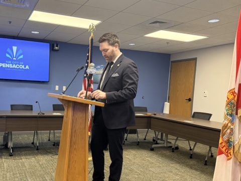 Man in a dark suit speaking at a wooden podium in a conference room, with a Pensacola city screen in the background and an American flag nearby.