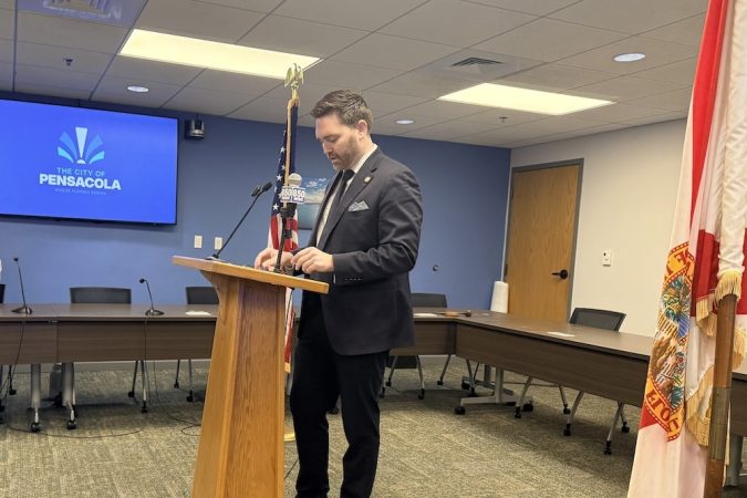 Man in a dark suit speaking at a wooden podium in a conference room, with a Pensacola city screen in the background and an American flag nearby.