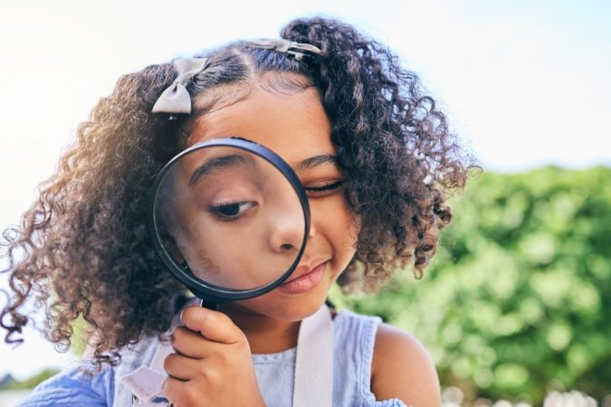 Girl outdoors holding a magnifying glass to her eye and peering through it playfully.