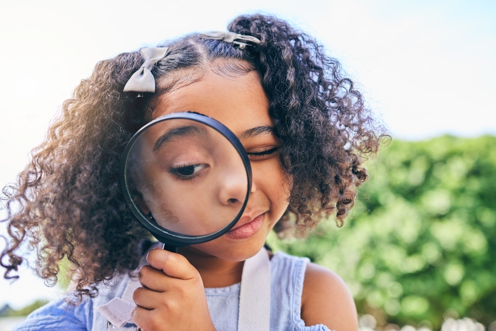 Girl outdoors holding a magnifying glass to her eye and peering through it playfully.