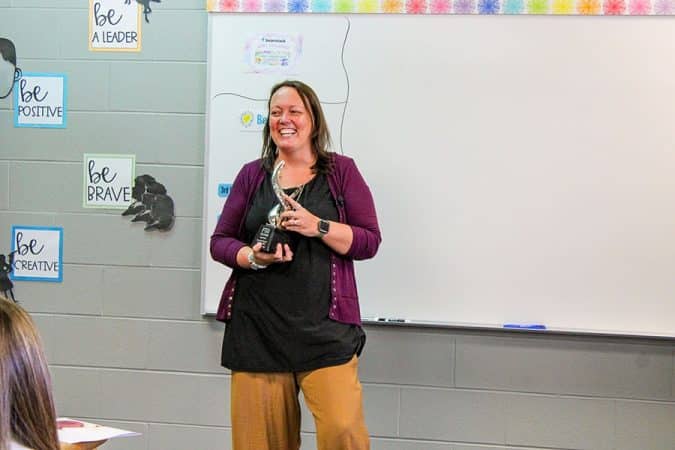 Smiling woman in a purple cardigan holds an award in a classroom setting with be-positive posters on the wall behind her.