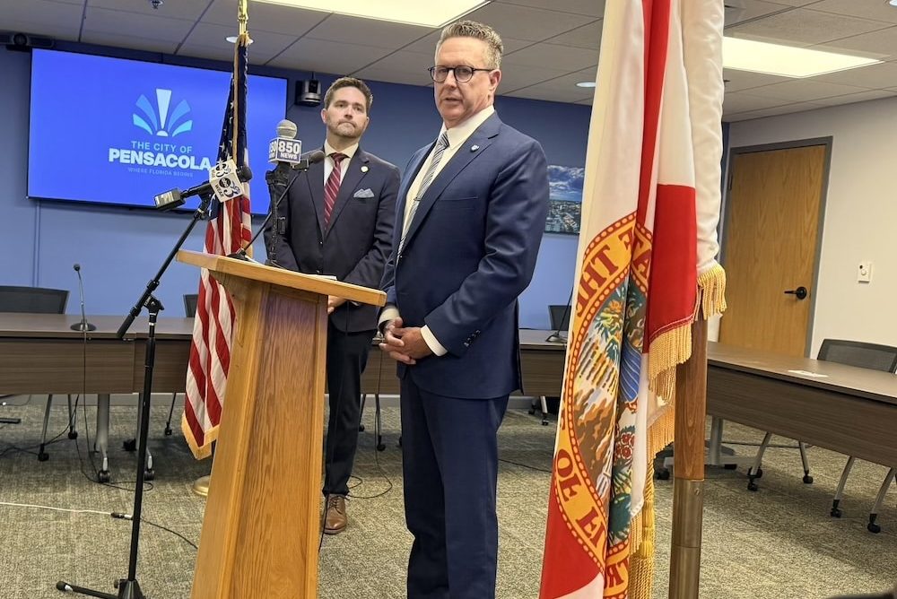 Two men in suits stand at a wooden podium during a press conference in a government meeting room, with American and state flags nearby and a 'City of Pensacola' backdrop.