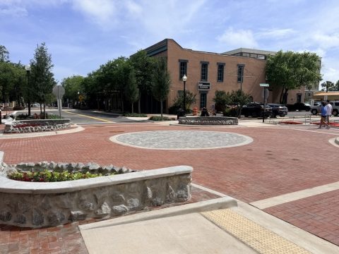 Brick-paved town square with a circular stone centerpiece, low stone planters, and trees, against a brick building in the background on a sunny day.