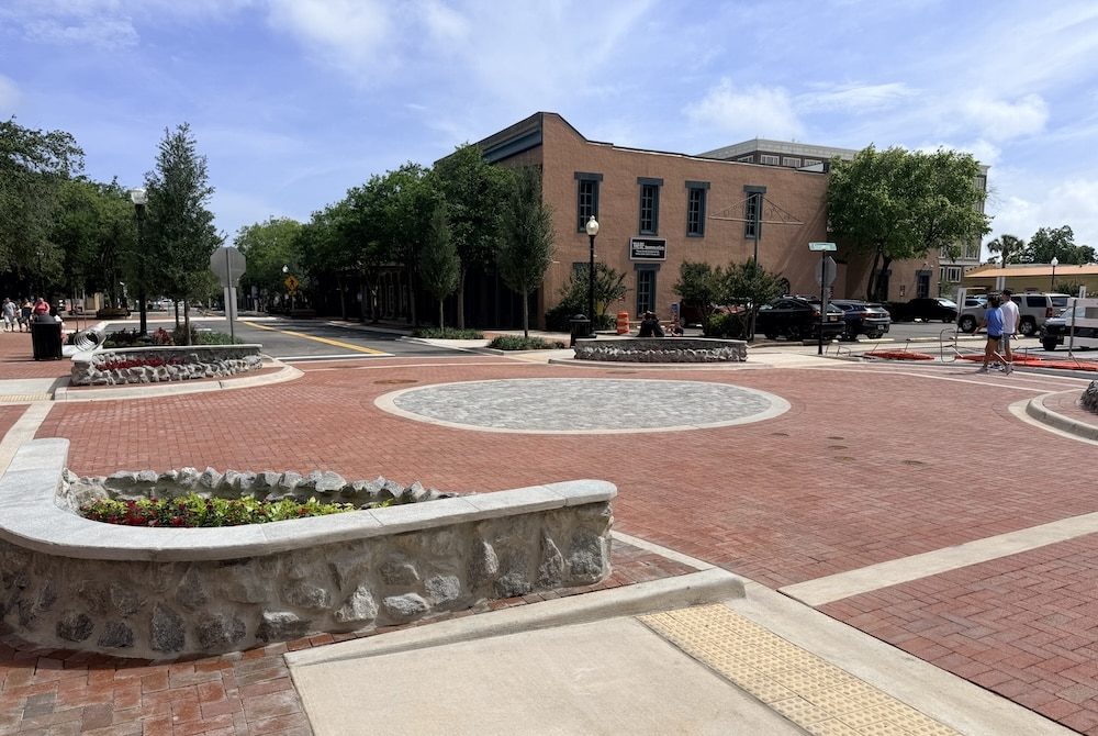 Brick-paved town square with a circular stone centerpiece, low stone planters, and trees, against a brick building in the background on a sunny day.