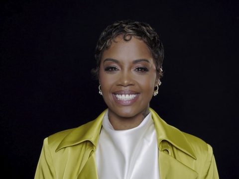 Smiling woman with short curls wearing a bright yellow jacket over a white top, against a black background.