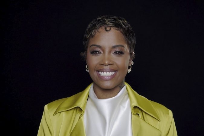Smiling woman with short curls wearing a bright yellow jacket over a white top, against a black background.