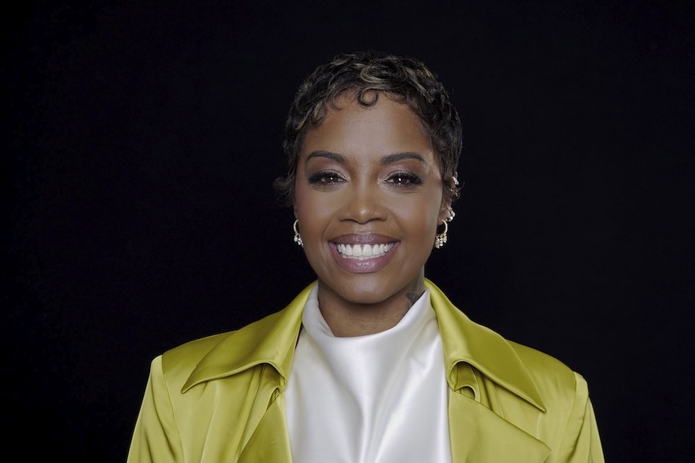 Smiling woman with short curls wearing a bright yellow jacket over a white top, against a black background.