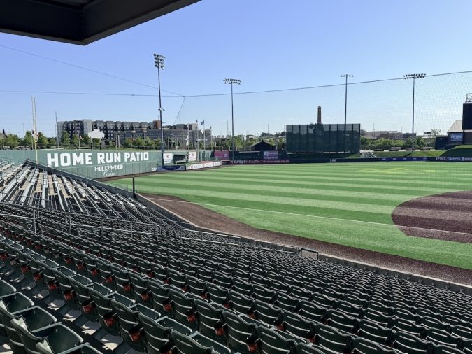 Empty stadium seating overlooking a baseball field with a large 'HOME RUN PATIO' sign along the outfield wall under a clear blue sky.