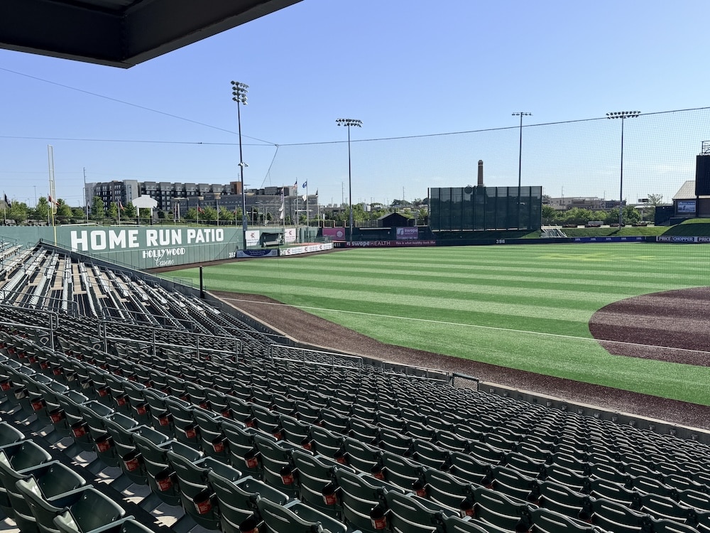 Empty stadium seating overlooking a baseball field with a large 'HOME RUN PATIO' sign along the outfield wall under a clear blue sky.