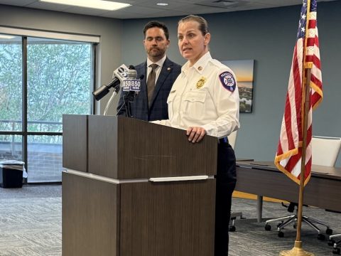 Police officer at a podium giving a press briefing, with a man in a suit behind her and an American flag to the right.