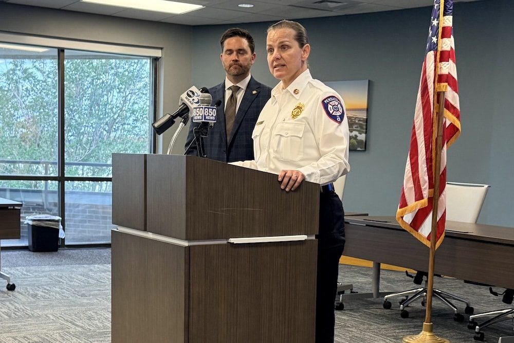 Police officer at a podium giving a press briefing, with a man in a suit behind her and an American flag to the right.