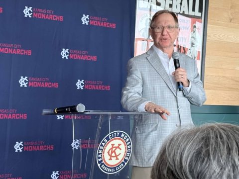 Man in a light gray suit speaks into a handheld microphone at a clear podium with Kansas City Monarchs branding in the background.