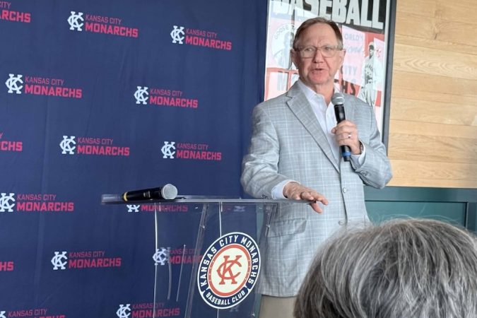 Man in a light gray suit speaks into a handheld microphone at a clear podium with Kansas City Monarchs branding in the background.