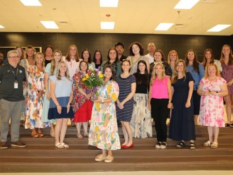 Group of about 25 people posing on indoor venue steps, with a woman in a colorful dress at the center holding a bouquet of flowers while others smile around her.