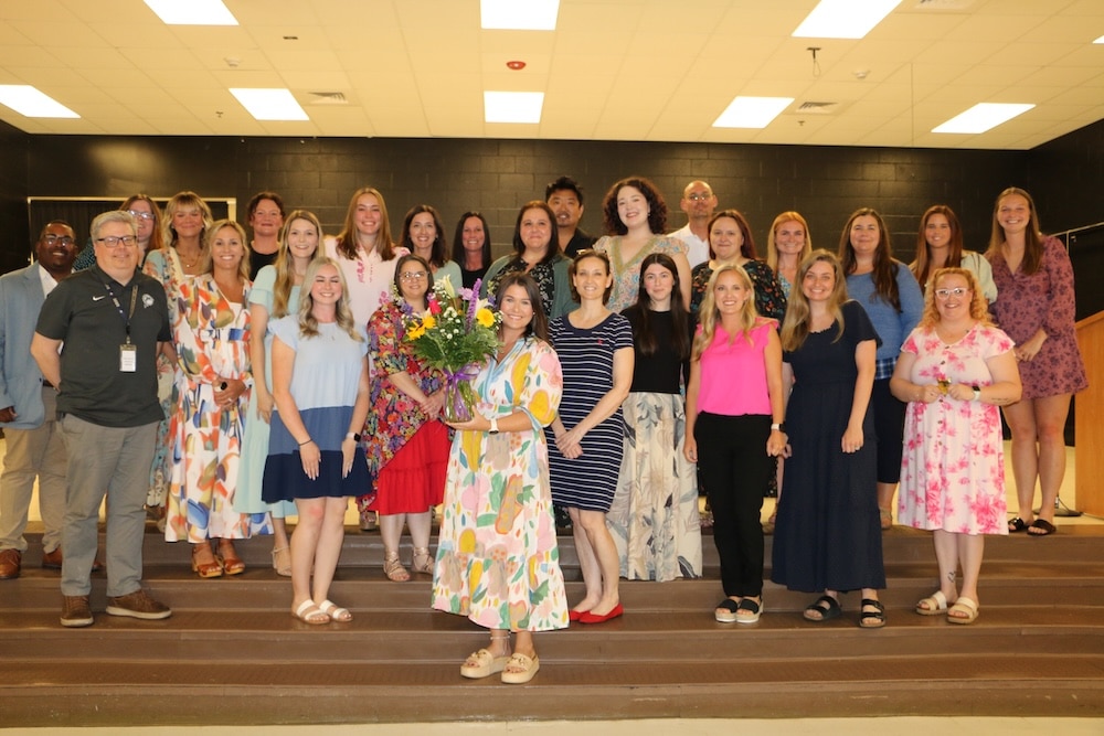 Group of about 25 people posing on indoor venue steps, with a woman in a colorful dress at the center holding a bouquet of flowers while others smile around her.
