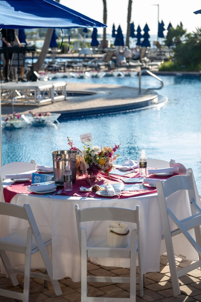 Poolside dining table with a pink runner, floral centerpiece, and water bottles, by a sunny resort pool with blue umbrellas in the background