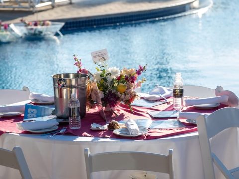 Poolside dining table with a pink runner, floral centerpiece, and water bottles, by a sunny resort pool with blue umbrellas in the background