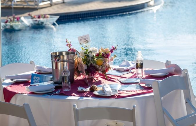 Poolside dining table with a pink runner, floral centerpiece, and water bottles, by a sunny resort pool with blue umbrellas in the background