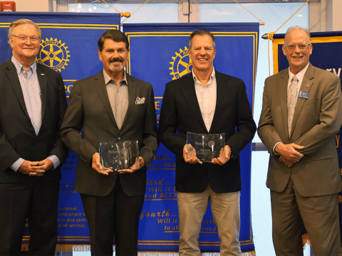 Four men in suits pose at a Rotary event; two middle men hold glass awards, blue Rotary banners behind them.