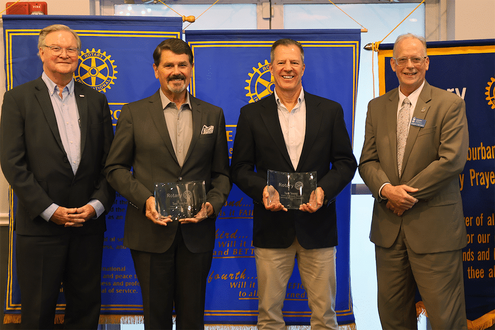 Four men in suits pose at a Rotary event; two middle men hold glass awards, blue Rotary banners behind them.