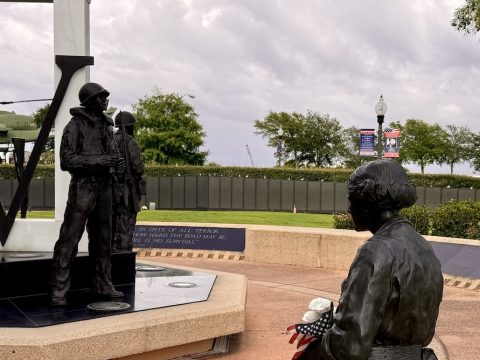 Bronze statues of soldiers at a war memorial, one standing with a rifle and another seated, with an American flag in view and a helicopter monument in the background.