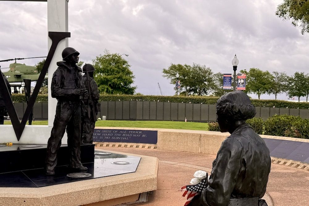 Bronze statues of soldiers at a war memorial, one standing with a rifle and another seated, with an American flag in view and a helicopter monument in the background.