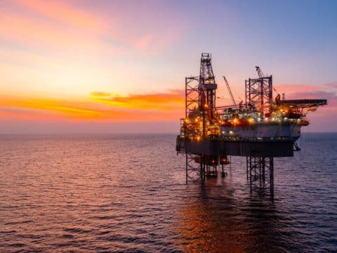 Offshore oil rig lit by lights at sunset on calm ocean sea.
