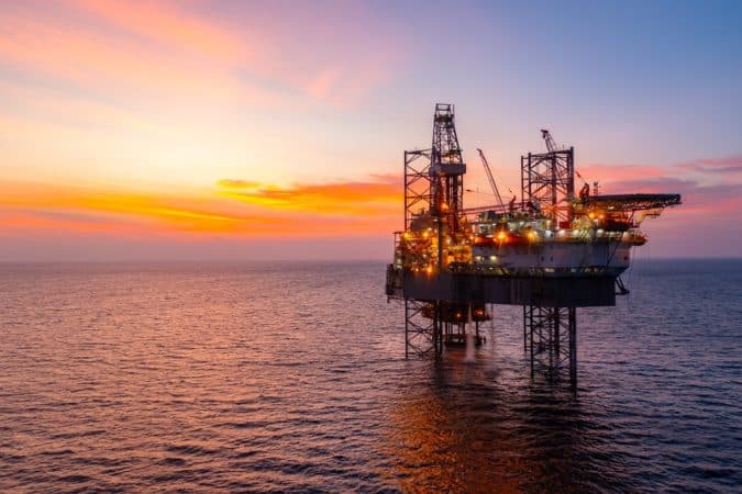 Offshore oil rig lit by lights at sunset on calm ocean sea.