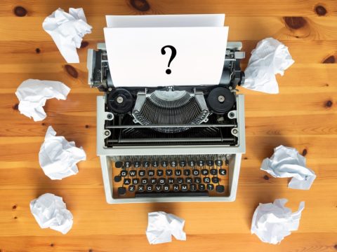 Vintage typewriter on a wooden desk with a sheet showing a large question mark, surrounded by crumpled papers, symbolizing writer's block or brainstorming.