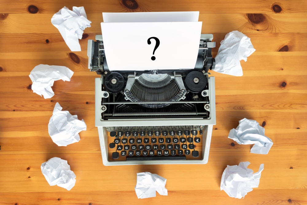 Vintage typewriter on a wooden desk with a sheet showing a large question mark, surrounded by crumpled papers, symbolizing writer's block or brainstorming.