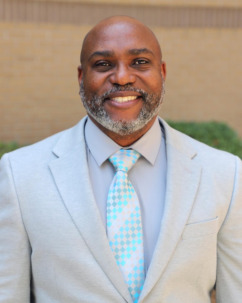 Smiling Black man in a light gray suit with a blue-checkered tie, standing outdoors.
