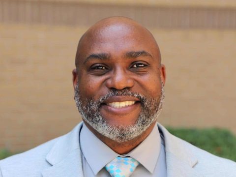 Smiling Black man in a light gray suit with a blue-checkered tie, standing outdoors.