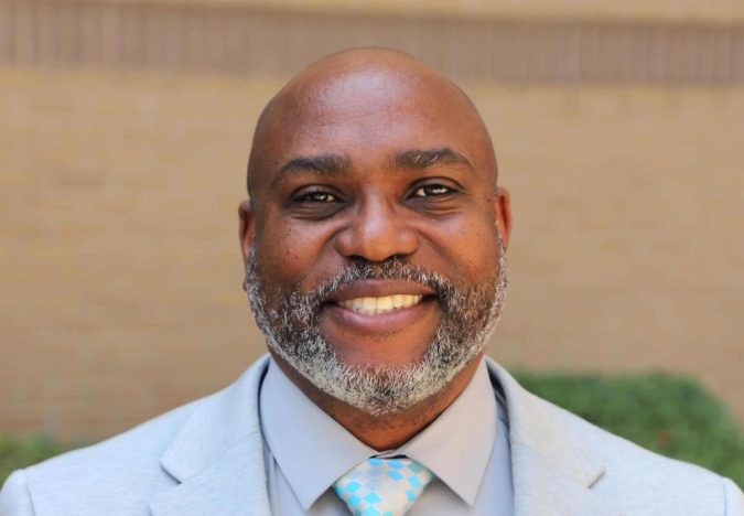 Smiling Black man in a light gray suit with a blue-checkered tie, standing outdoors.