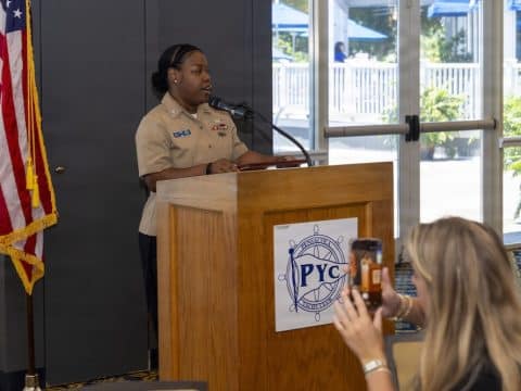Black woman in a tan military-style uniform speaks at a wooden podium with a PYC emblem, American flag to her left, as someone records with a smartphone in the foreground.