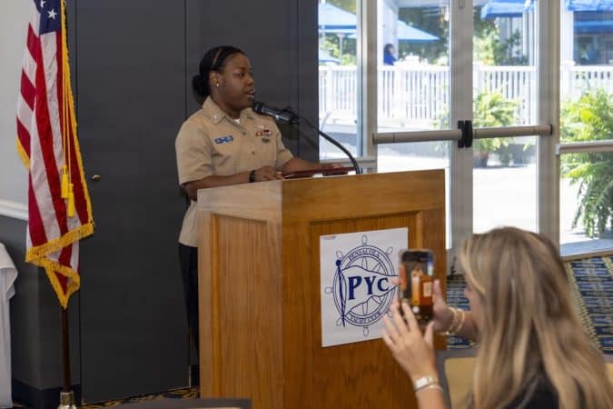 Black woman in a tan military-style uniform speaks at a wooden podium with a PYC emblem, American flag to her left, as someone records with a smartphone in the foreground.