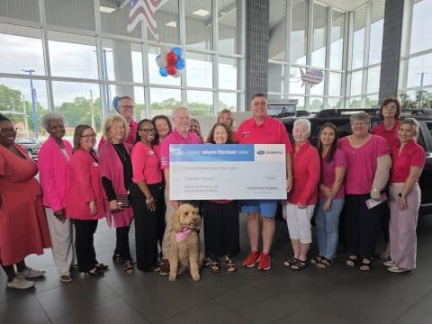 Group of adults in pink clothing posing with a large charity check inside a car dealership showroom, with a pink dog in front wearing a scarf and balloons overhead.