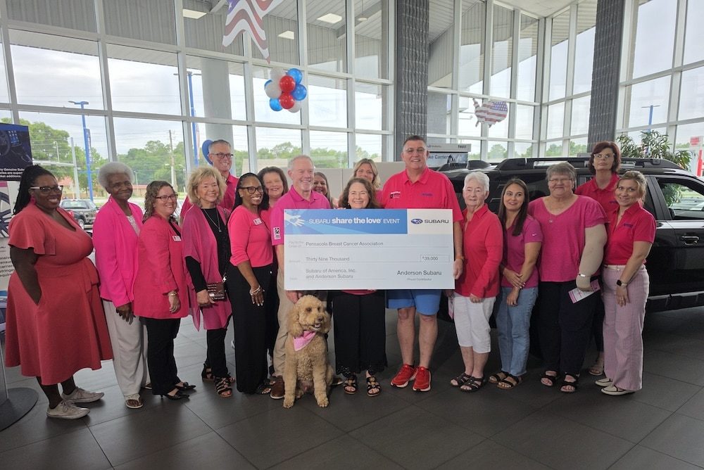 Group of adults in pink clothing posing with a large charity check inside a car dealership showroom, with a pink dog in front wearing a scarf and balloons overhead.