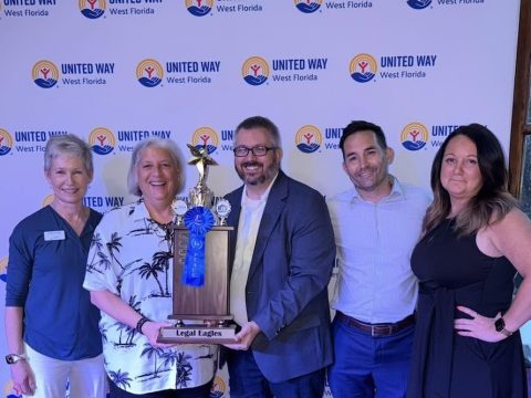 Five adults pose with a large trophy reading 'Legal Eagles' in front of a United Way West Florida backdrop.